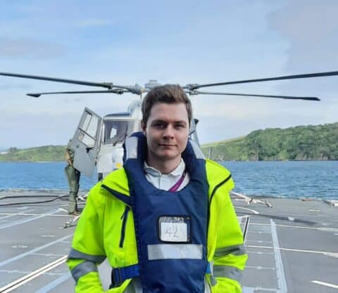 Male Maritime Defence worker in a high visibility coat stands in front of a helicopter on a coastal helipad