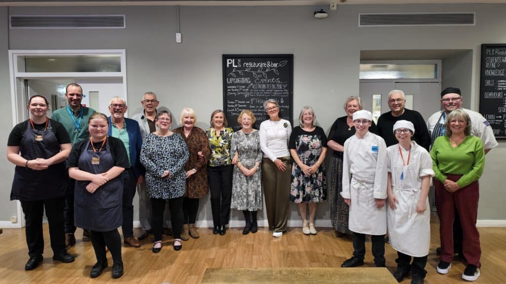 Fourteen members of the class of 1975-77 stand with current hospitality students in City College Plymouth's PL1 restaurant, everyone smiling