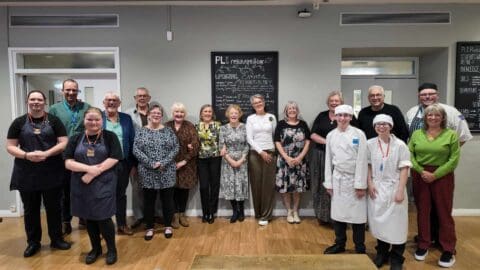 Fourteen members of the class of 1975-77 stand with current hospitality students in City College Plymouth's PL1 restaurant, everyone smiling