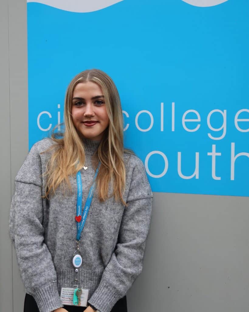 Female apprentice Lauren Jones wearing a lanyard leaning against a wall emblazoned with the light blue City College Plymouth logo.